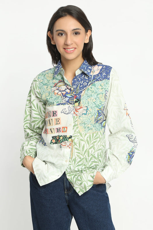 A woman modeling a long-sleeve button-up shirt with a multicolor patchwork print featuring green tropical leaves, blue floral details, and a white pocket with colorful text, paired with dark blue jeans against a plain white background.