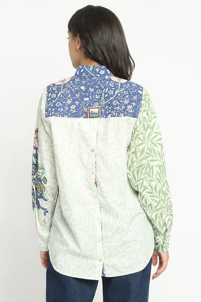 A woman shown from the back wearing a long-sleeve button-up shirt with a patchwork design: a blue floral yoke, a light green leafy sleeve, and a pale cream body, paired with dark blue jeans against a plain white background.