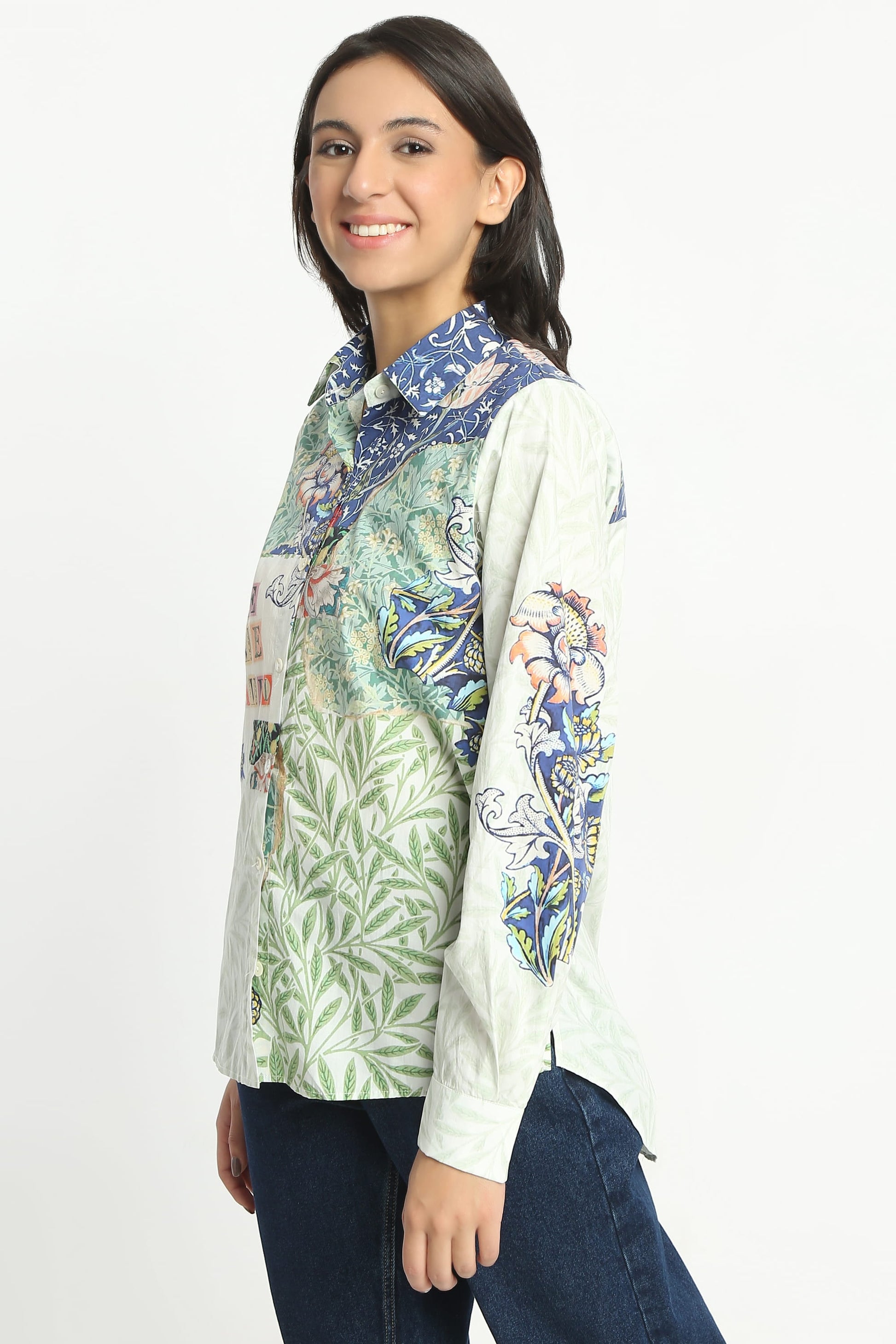 A woman modeling a long-sleeve button-up shirt with a mixed botanical and floral print in green, blue, and white, paired with dark blue jeans against a plain white background.