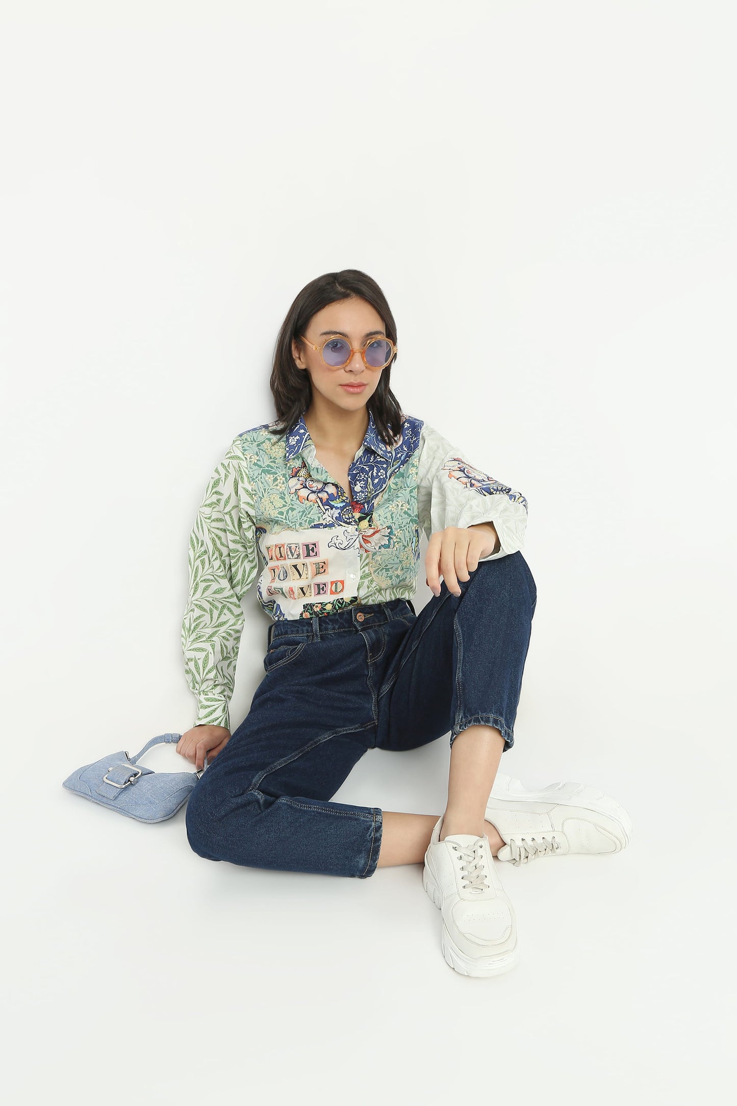 A model sits on a white studio floor wearing a light green and white patterned long-sleeve shirt with colorful graphic prints, dark blue jeans, white sneakers, and light blue sunglasses, holding a light blue handbag.