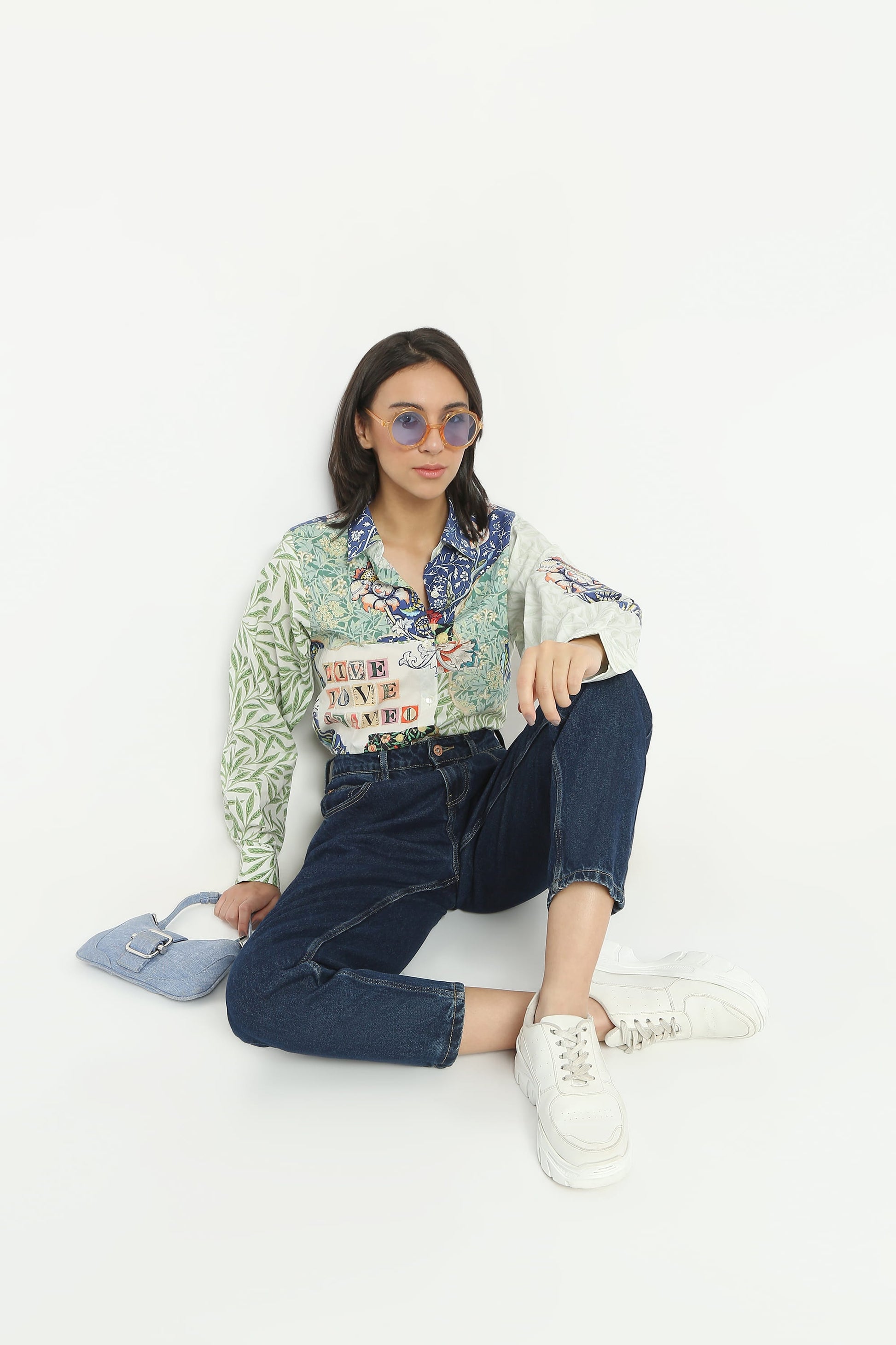 A model sits on a white studio floor wearing a light green and white patterned long-sleeve shirt with colorful graphic prints, dark blue jeans, white sneakers, and light blue sunglasses, holding a light blue handbag.