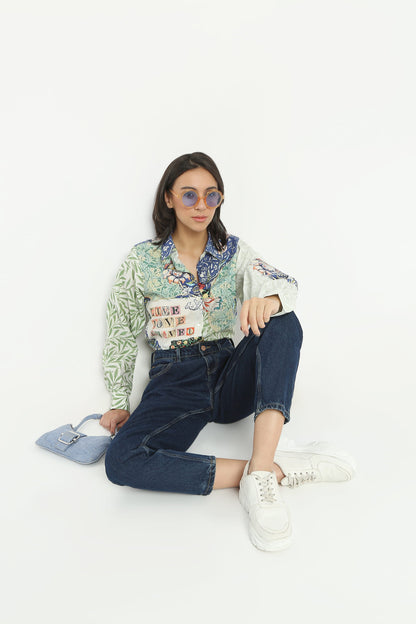 A model sits on a white studio floor wearing a light green and white patterned long-sleeve shirt with colorful graphic prints, dark blue jeans, white sneakers, and light blue sunglasses, holding a light blue handbag.
