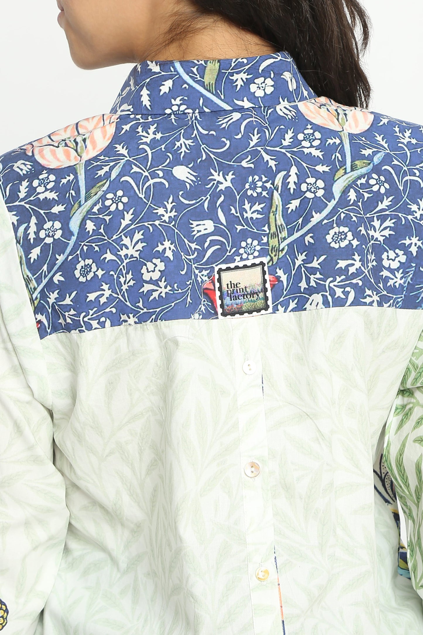 Close-up back view of a person wearing a collared button-up shirt with a blue floral yoke and an off-white textured body. A small square brand patch is visible near the collar.