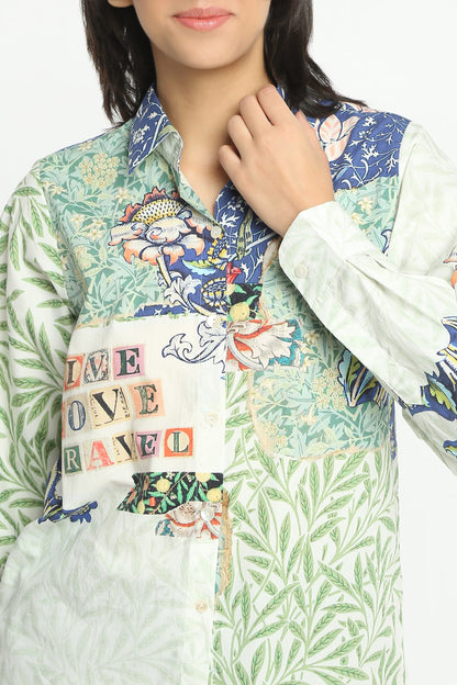 A close-up of a model wearing a long-sleeve button-up shirt with a multicolor patchwork print. The shirt features a white base with green leaf patterns, blue floral sections, and a central patch with the words 'LOVE TRAVELO' in colorful block letters.
