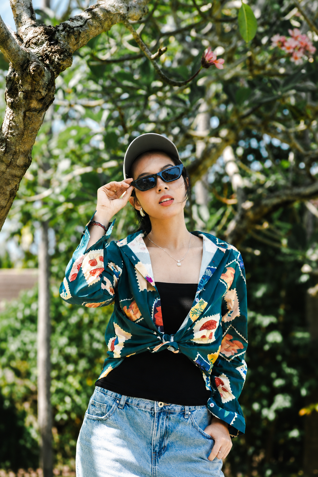 A woman wearing a dark teal patterned cardigan with a colorful floral and abstract print, tied at the waist over a black top and light blue jeans, standing outdoors in front of a tree with green leaves and flowers.