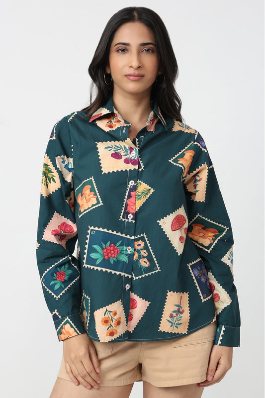 A woman modeling a dark teal button-up shirt with a colorful patchwork print of vintage-style illustrated cards, paired with beige shorts, against a light gray studio background.