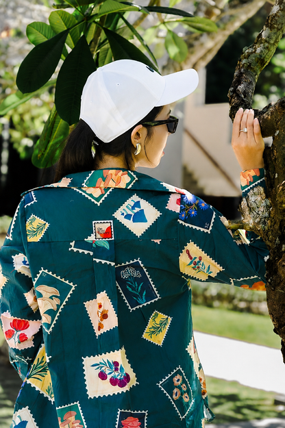 A woman wearing a dark teal button-up shirt with colorful patchwork floral prints, a white baseball cap, and black sunglasses, standing outdoors next to a tree trunk.