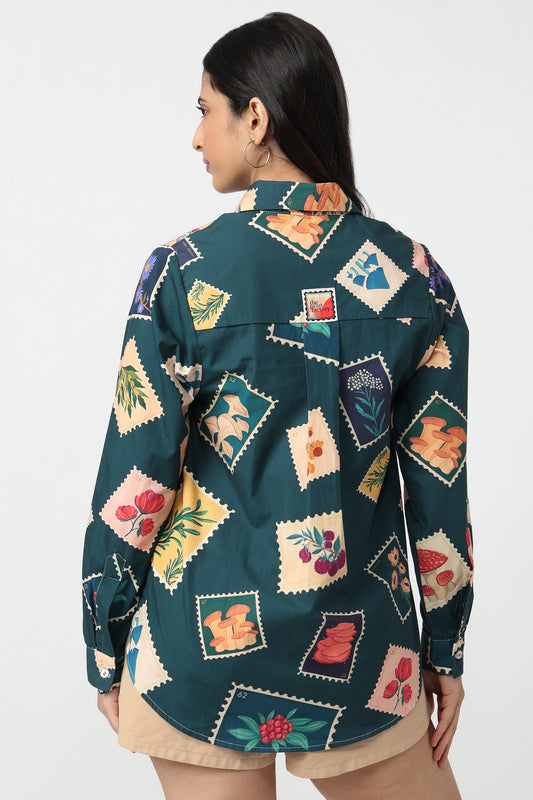 A woman shown from the back wearing a dark teal button-up shirt with a colorful patchwork-style print of vintage postage stamps and floral motifs, paired with beige shorts against a plain white studio background.