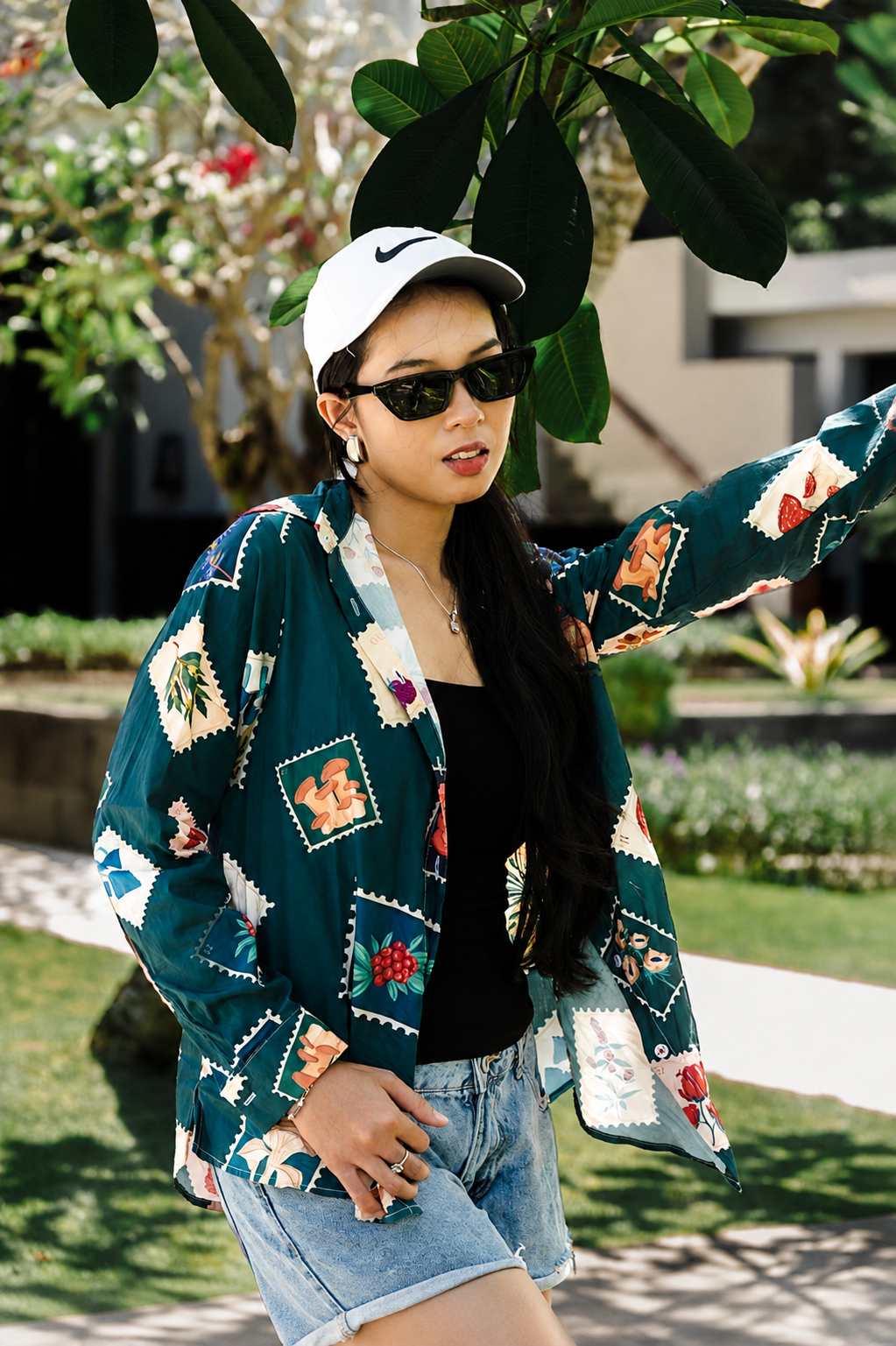 A woman wearing a dark teal green patchwork print cardigan, a white Nike cap, black sunglasses, and denim shorts, posing outdoors in a garden setting.