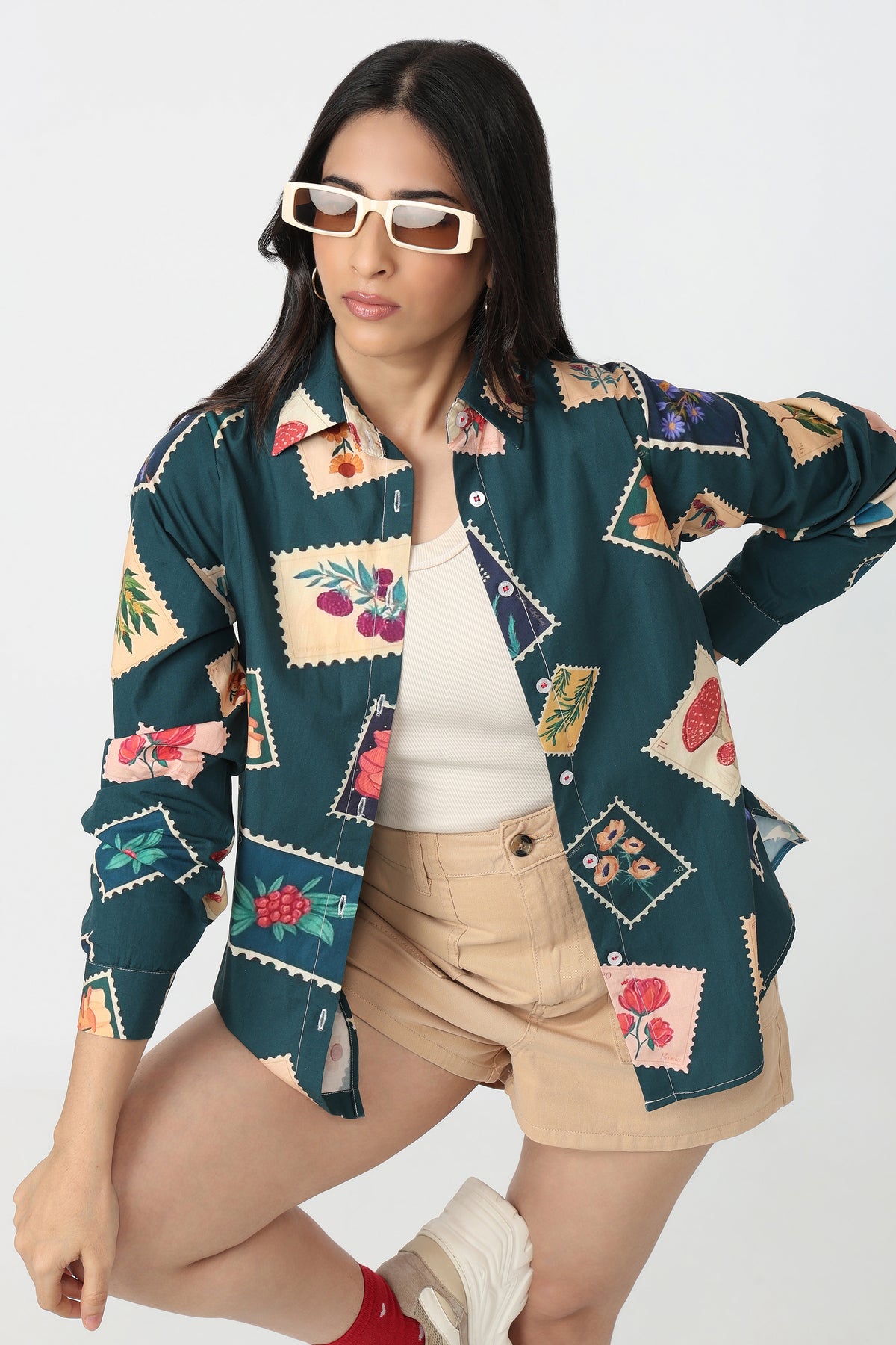 A woman modeling a dark teal button-up shirt with a colorful floral and patchwork print, styled with beige shorts, a white tank top, and white sneakers against a light gray background.