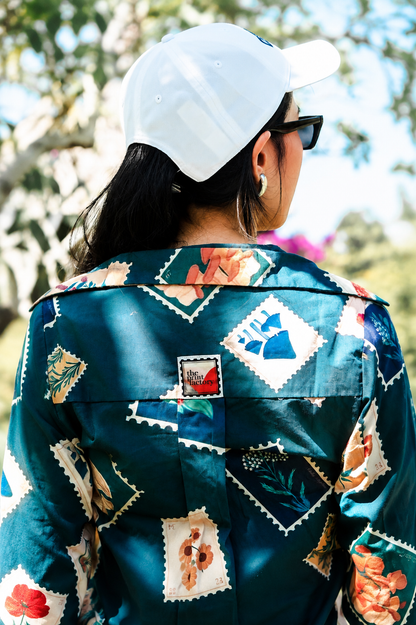 A person viewed from behind wearing a teal button-up shirt with a colorful patchwork print of vintage postage stamps, a white baseball cap, and black sunglasses in an outdoor setting.