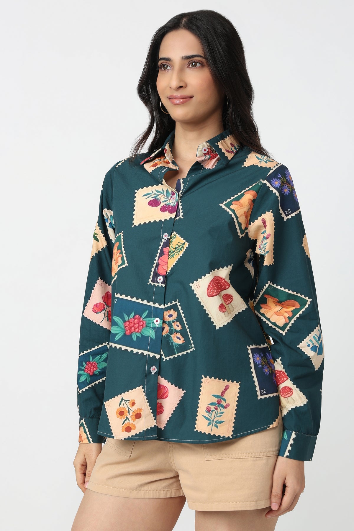 A woman wearing a dark teal long-sleeve button-up shirt with a colorful patchwork print of postage stamps, each containing different illustrated motifs like flowers, mushrooms, and fruit, paired with beige shorts against a plain white background.