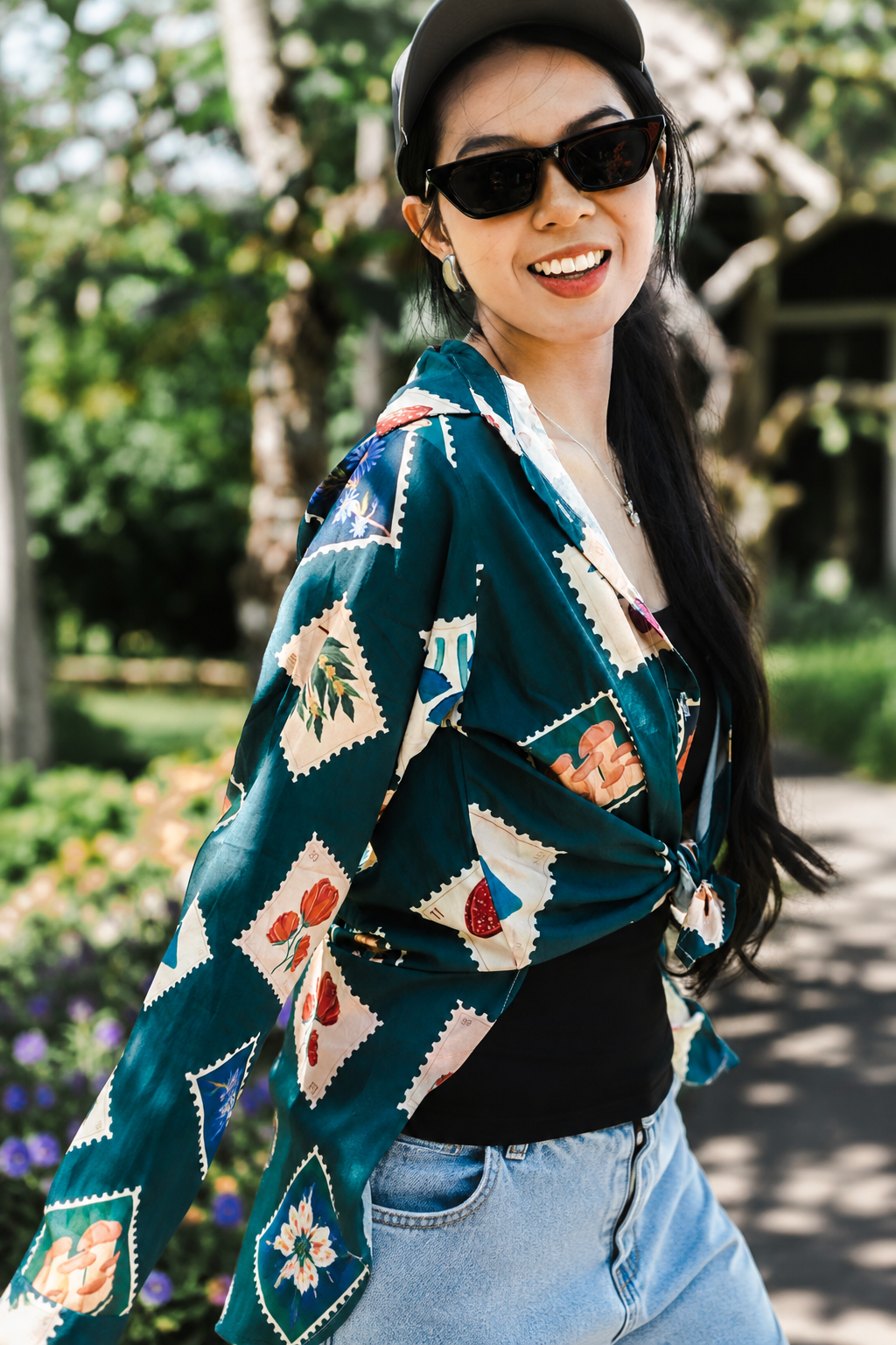 A woman wearing a dark teal patterned kimono-style cardigan over a black top and light blue jeans, standing outdoors in a garden setting.