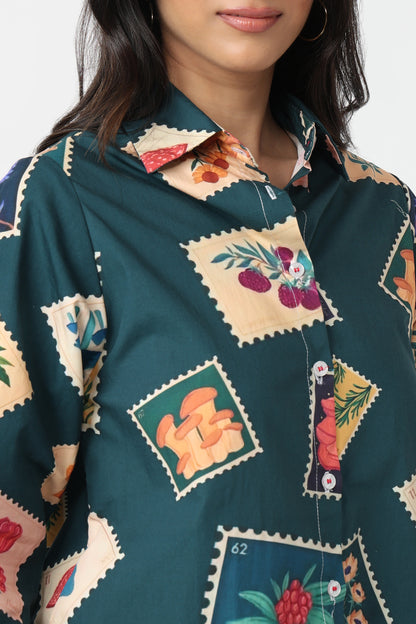A close-up of a woman wearing a dark teal button-up shirt with a colorful patchwork print of vintage postage stamps featuring botanical and abstract designs.