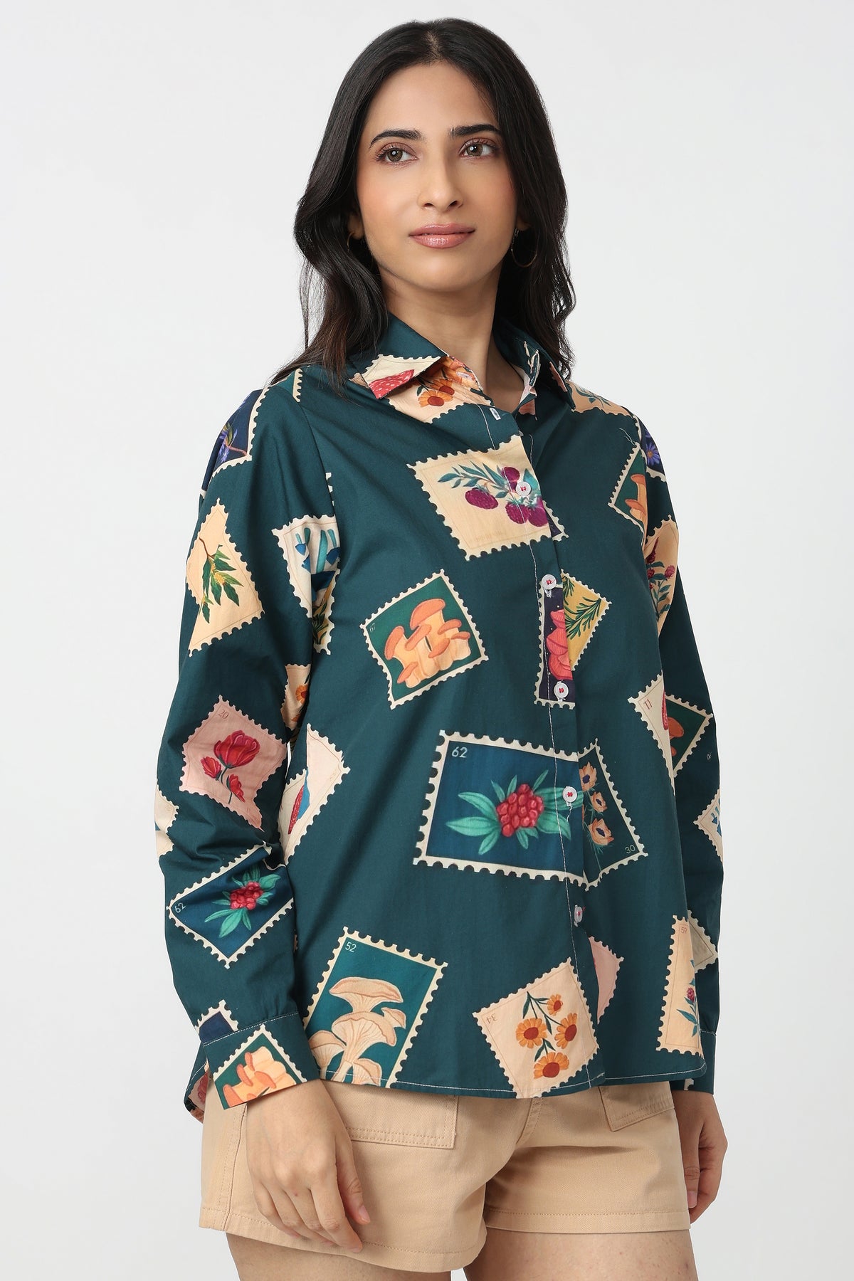 A woman modeling a dark teal long-sleeve button-up shirt with a colorful patchwork floral print, paired with beige shorts, against a plain light gray background.