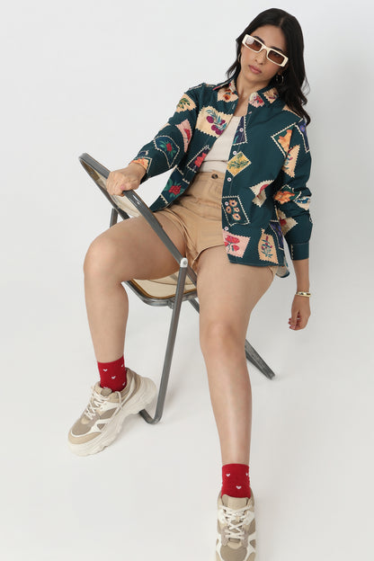A woman models a dark teal button-up shirt with a colorful geometric patchwork print, paired with beige shorts, red socks, and chunky sneakers while sitting on a folding chair against a white studio background.