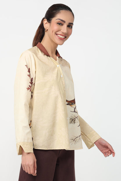 A woman modeling a cream-colored long-sleeve button-up shirt with brown collar trim and embroidered details, paired with dark brown trousers, against a plain white studio background.