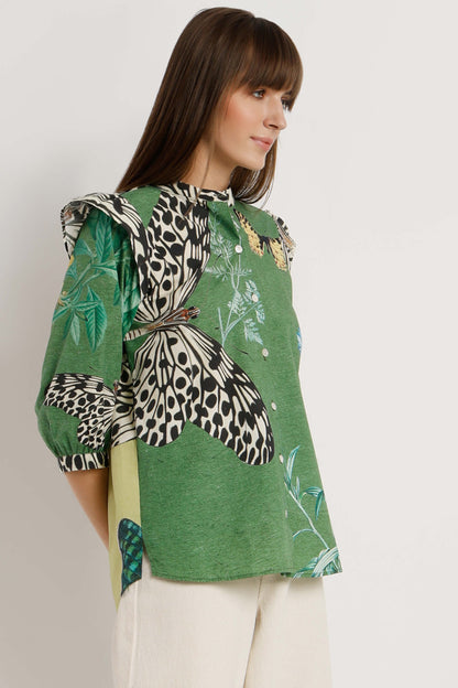 A woman wearing a green blouse with a large butterfly print, leopard print sleeve, and white pants, photographed against a plain light background.