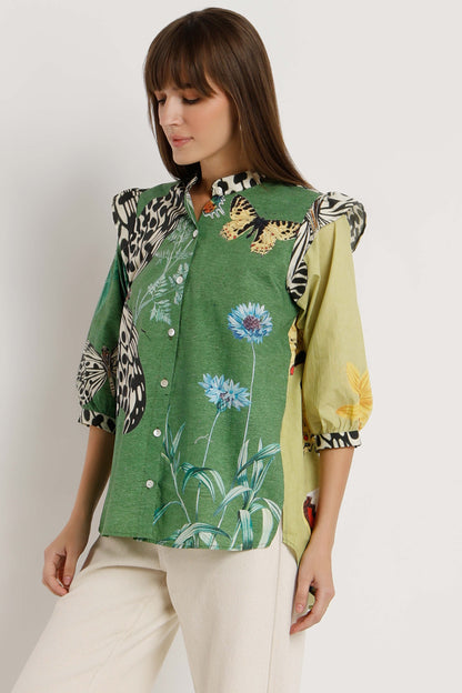 A woman wearing a green patchwork button-up blouse with butterfly and floral prints, paired with cream pants, photographed against a plain light background.