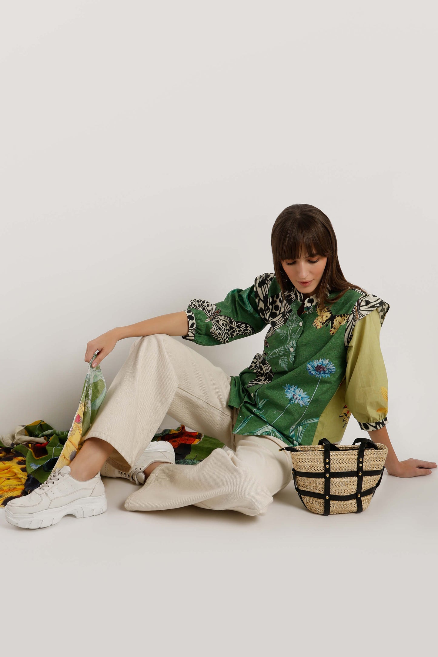 A model sits on a white floor wearing a green floral and animal print shirt, cream wide-leg trousers, and white sneakers, next to a woven straw bag with black straps.
