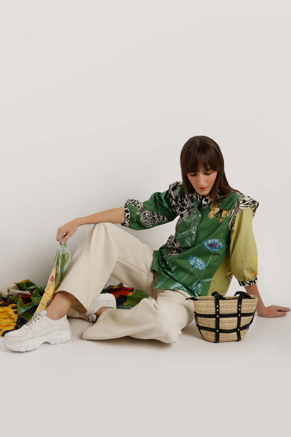 A model sits on a white floor wearing a green floral and animal print shirt, cream wide-leg trousers, and white sneakers, next to a woven straw bag with black straps.