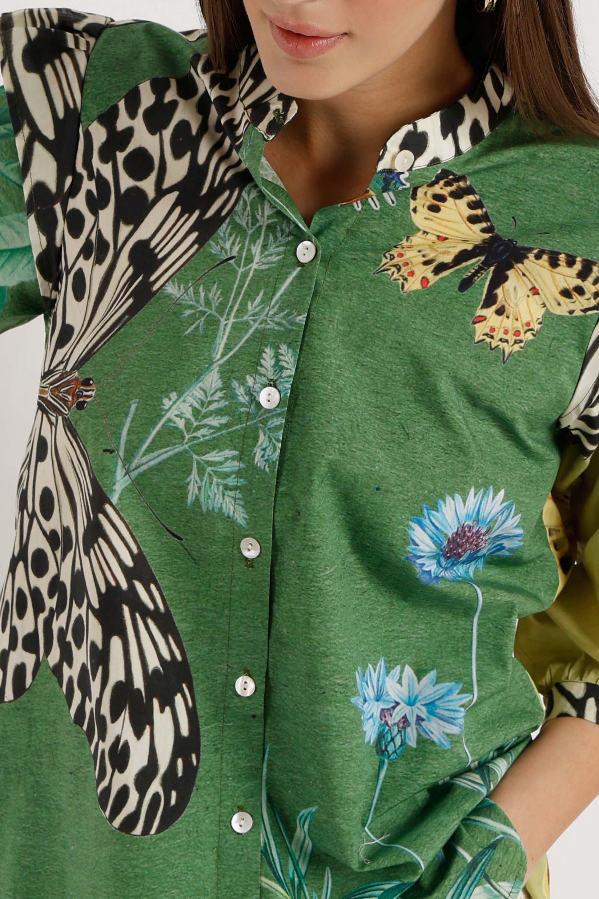 A close-up of a green button-front shirt with butterfly and floral prints, worn by a model. The shirt features a large black-and-white spotted butterfly motif on the left side and a yellow-and-black butterfly on the right, alongside blue flowers.