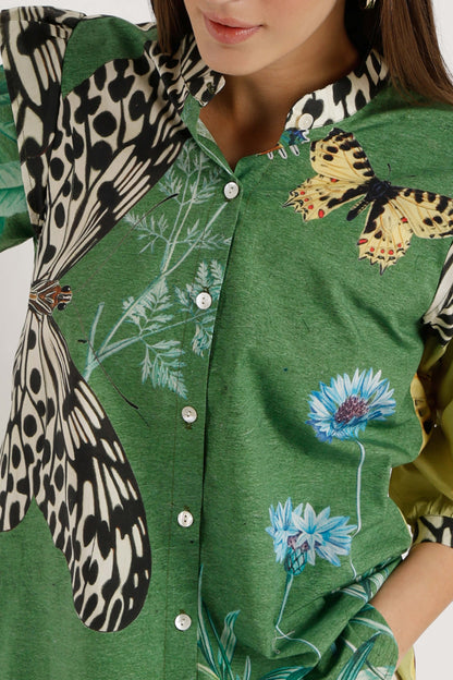 A close-up of a green button-front shirt with butterfly and floral prints, worn by a model. The shirt features a large black-and-white spotted butterfly motif on the left side and a yellow-and-black butterfly on the right, alongside blue flowers.