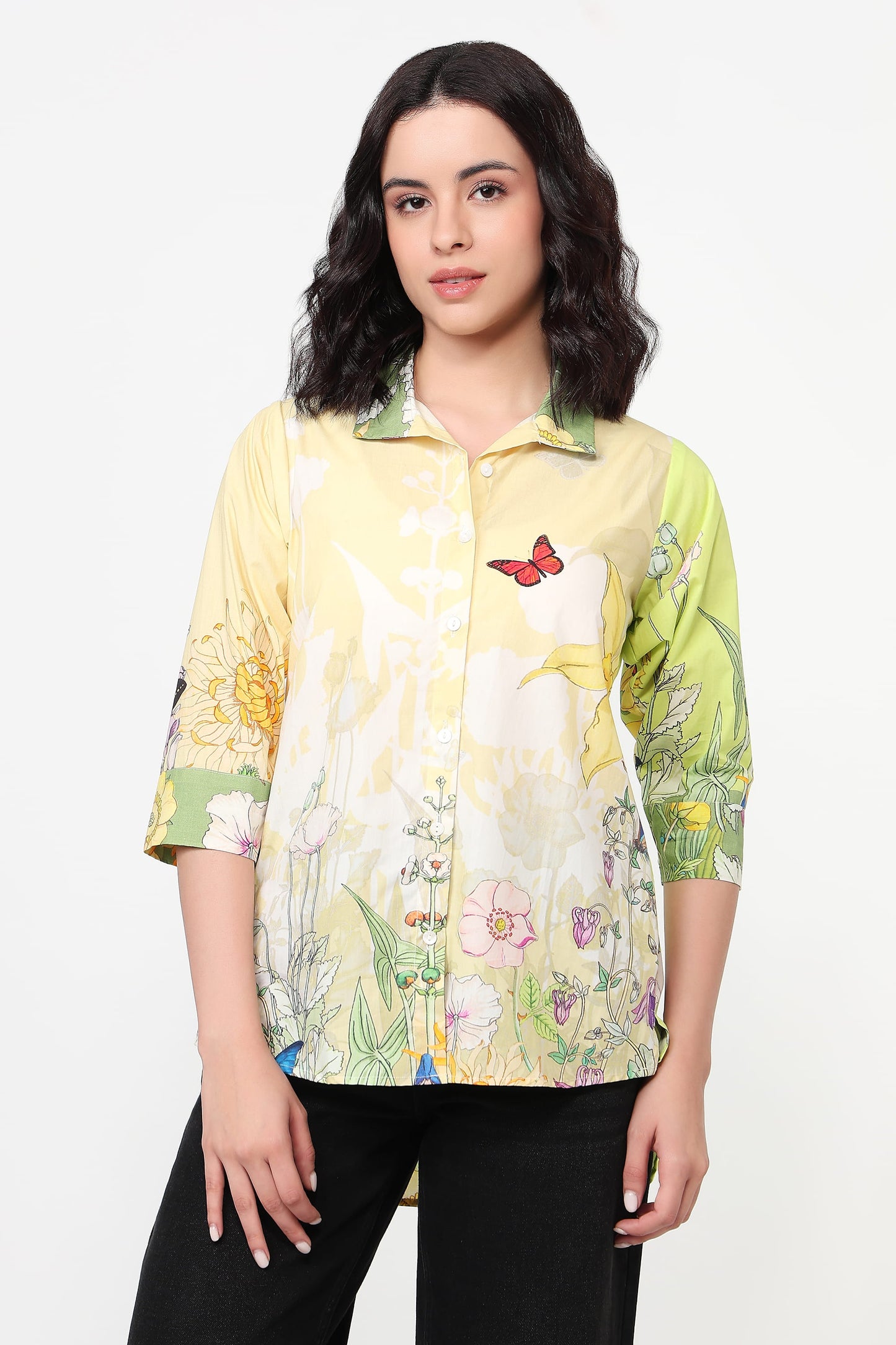 A woman modeling a short-sleeved button-up shirt with a yellow and green floral print, featuring a butterfly motif on the chest, paired with black pants against a plain white background.