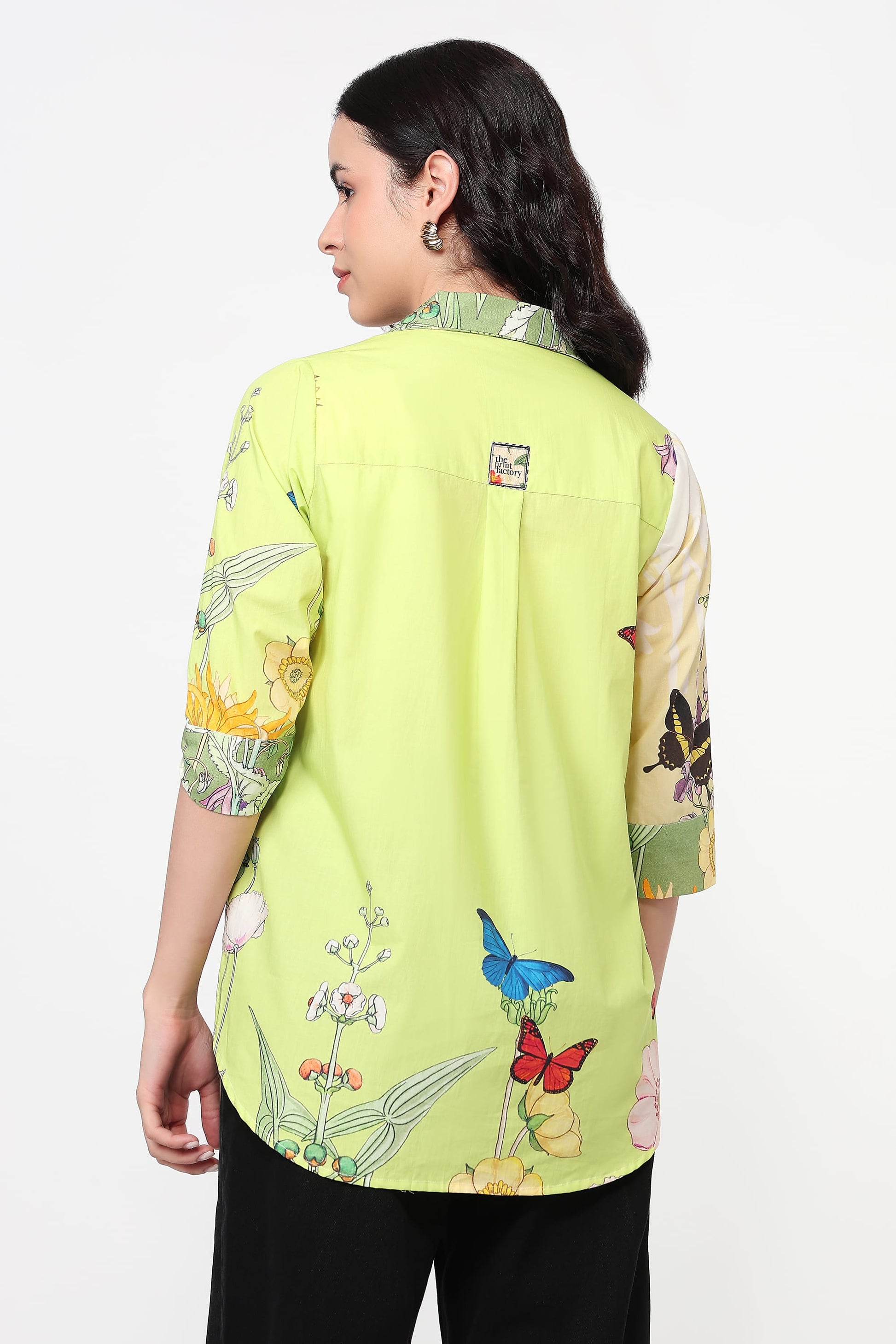 A woman shown from the back wearing a lime green shirt with colorful butterfly and floral prints on the sleeves and lower back, paired with black pants, against a plain white background.