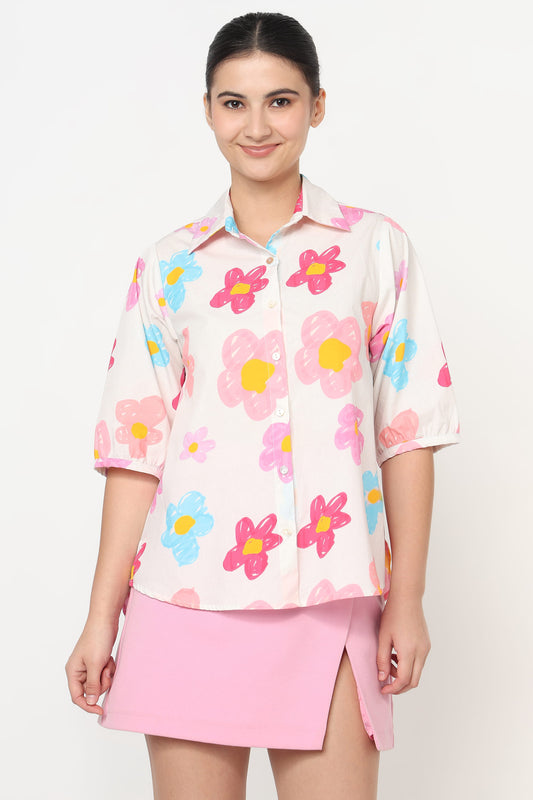A woman modeling a short-sleeve collared button-up shirt with a colorful floral print in pink, blue, and yellow on a light cream background, paired with a pink skirt, against a plain light gray studio background.