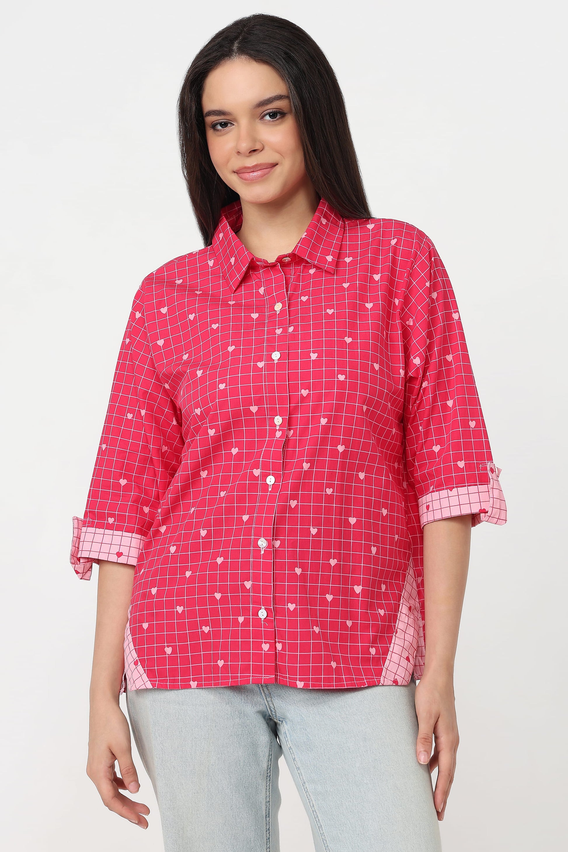 A woman wearing a pink and red checkered button-up shirt with small white heart patterns, paired with light gray jeans, against a plain white background.
