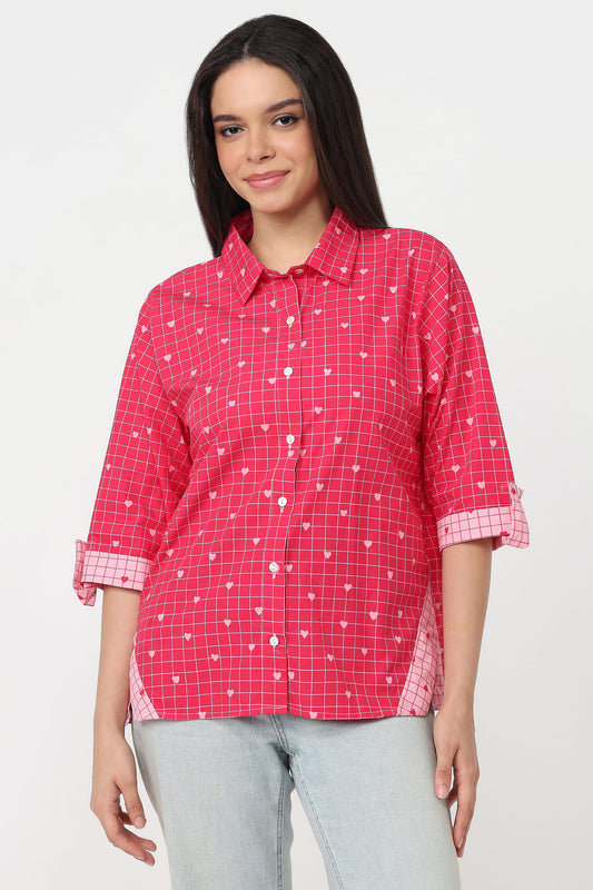 A woman wearing a pink and red checkered button-up shirt with small white heart patterns, paired with light gray jeans, against a plain white background.