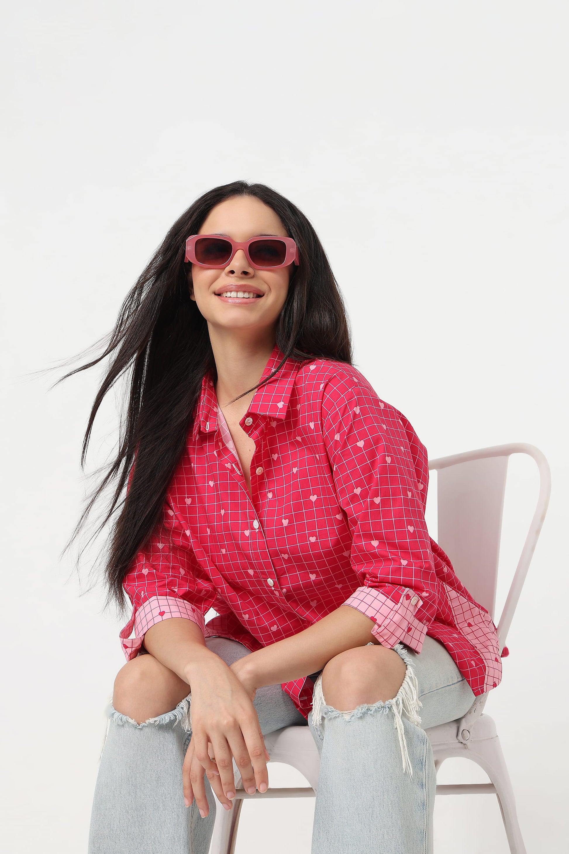 A woman with long black hair sits on a white chair wearing a pink and white checkered button-up shirt, light-wash distressed jeans, and pink sunglasses against a plain white background.
