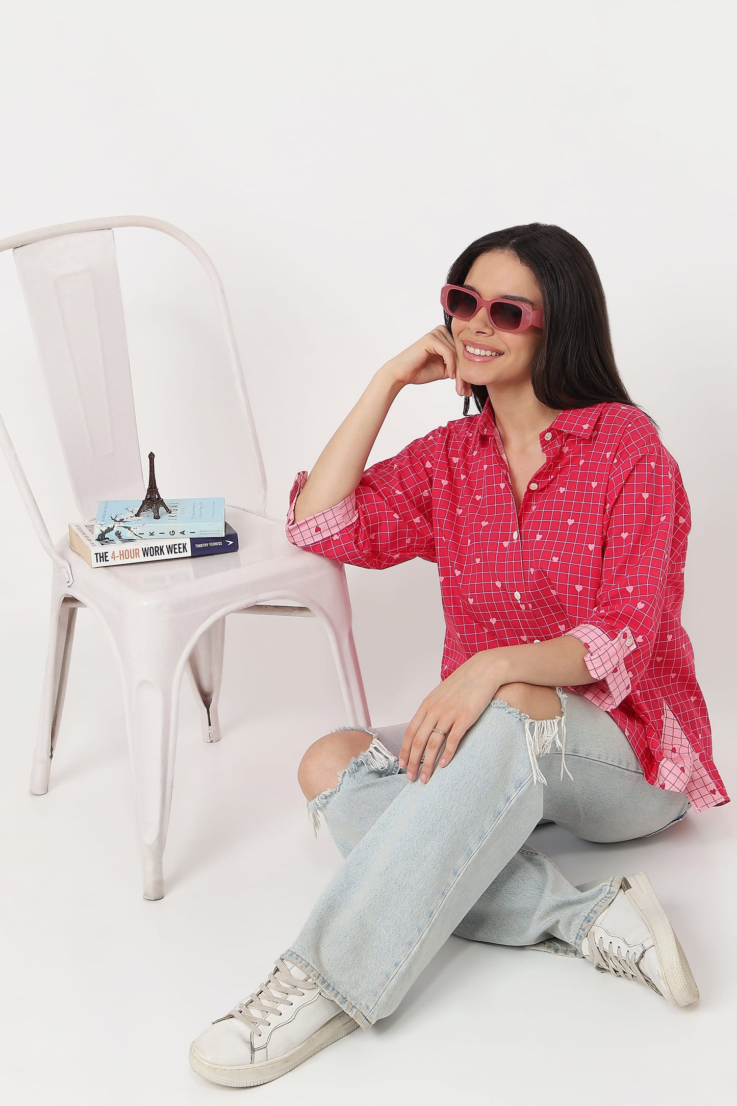 A woman wearing a pink and white checkered button-up shirt, light-wash ripped jeans, white sneakers, and pink sunglasses sits on a white floor next to a white metal chair with books.