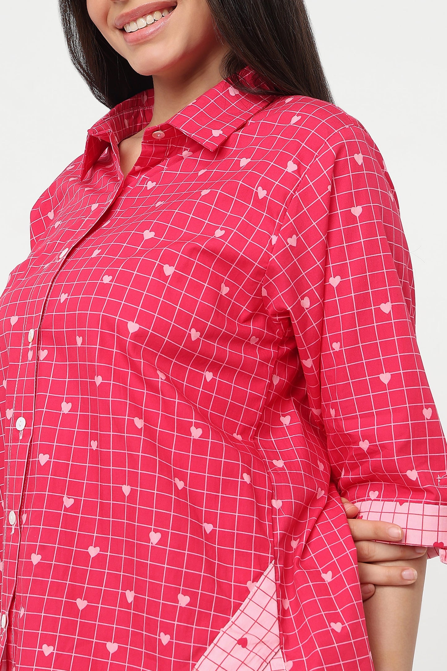 A close-up of a woman wearing a bright pink button-up shirt with a white grid pattern and small white heart motifs.