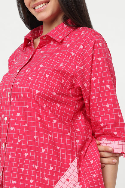 A close-up of a woman wearing a bright pink button-up shirt with a white grid pattern and small white heart motifs.