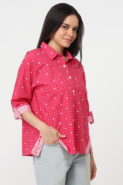 A woman wearing a pink and white checkered button-up shirt with rolled-up sleeves and light gray jeans, photographed against a plain light gray studio background.