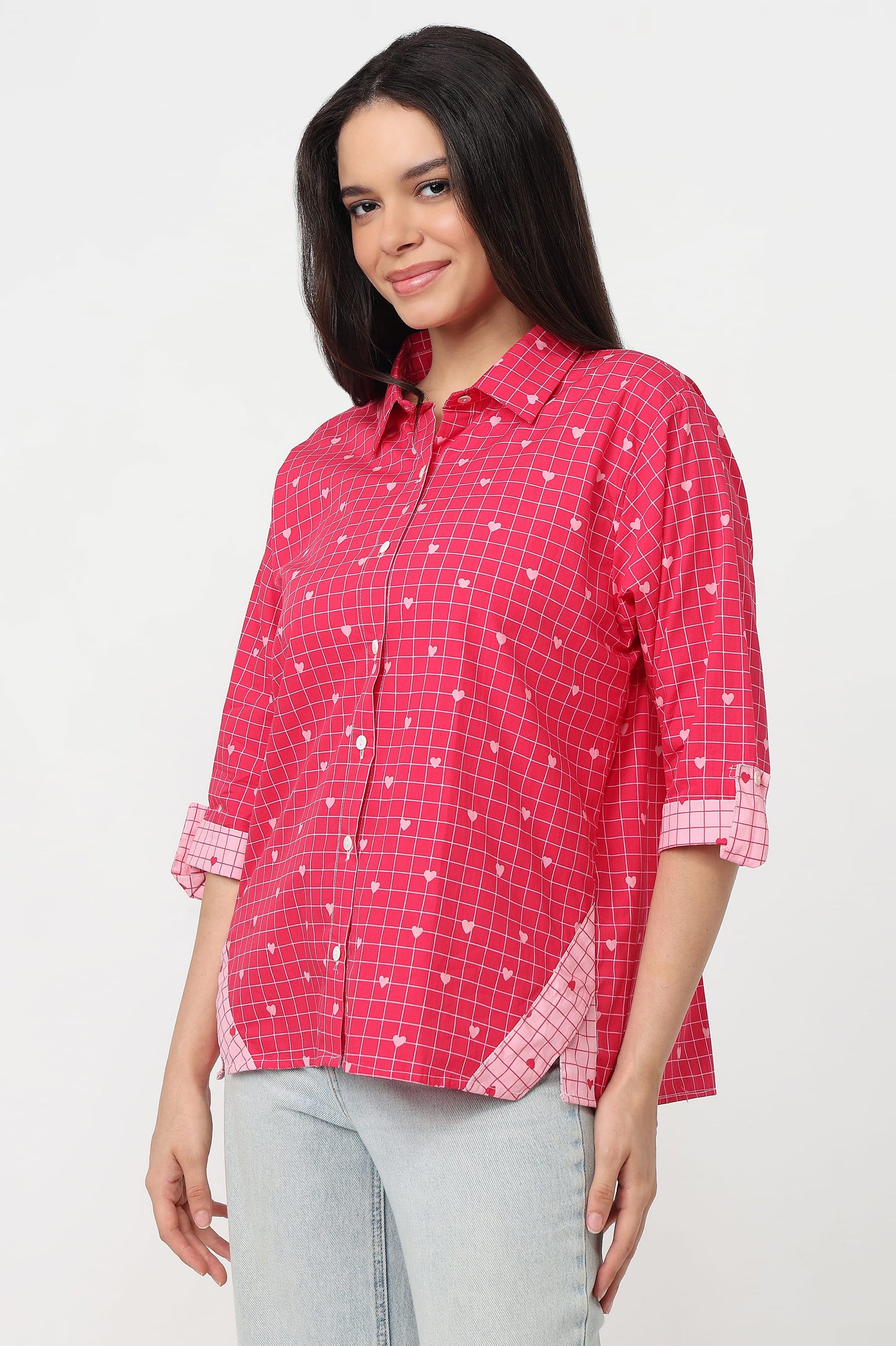 A woman wearing a pink checkered button-up shirt with white heart patterns, paired with light gray jeans, against a plain white background.