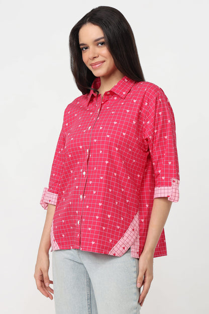 A woman wearing a pink checkered button-up shirt with white heart patterns, paired with light gray jeans, against a plain white background.