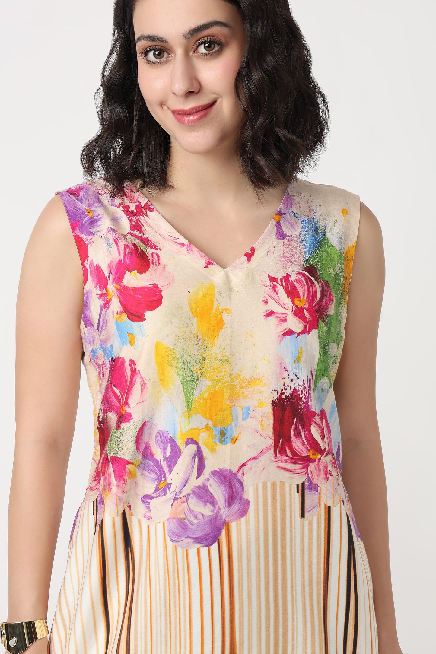 A woman wearing a sleeveless V-neck dress with a colourful floral print and vertical stripes on the bodice and, paired with black heels, against a plain light grey background.