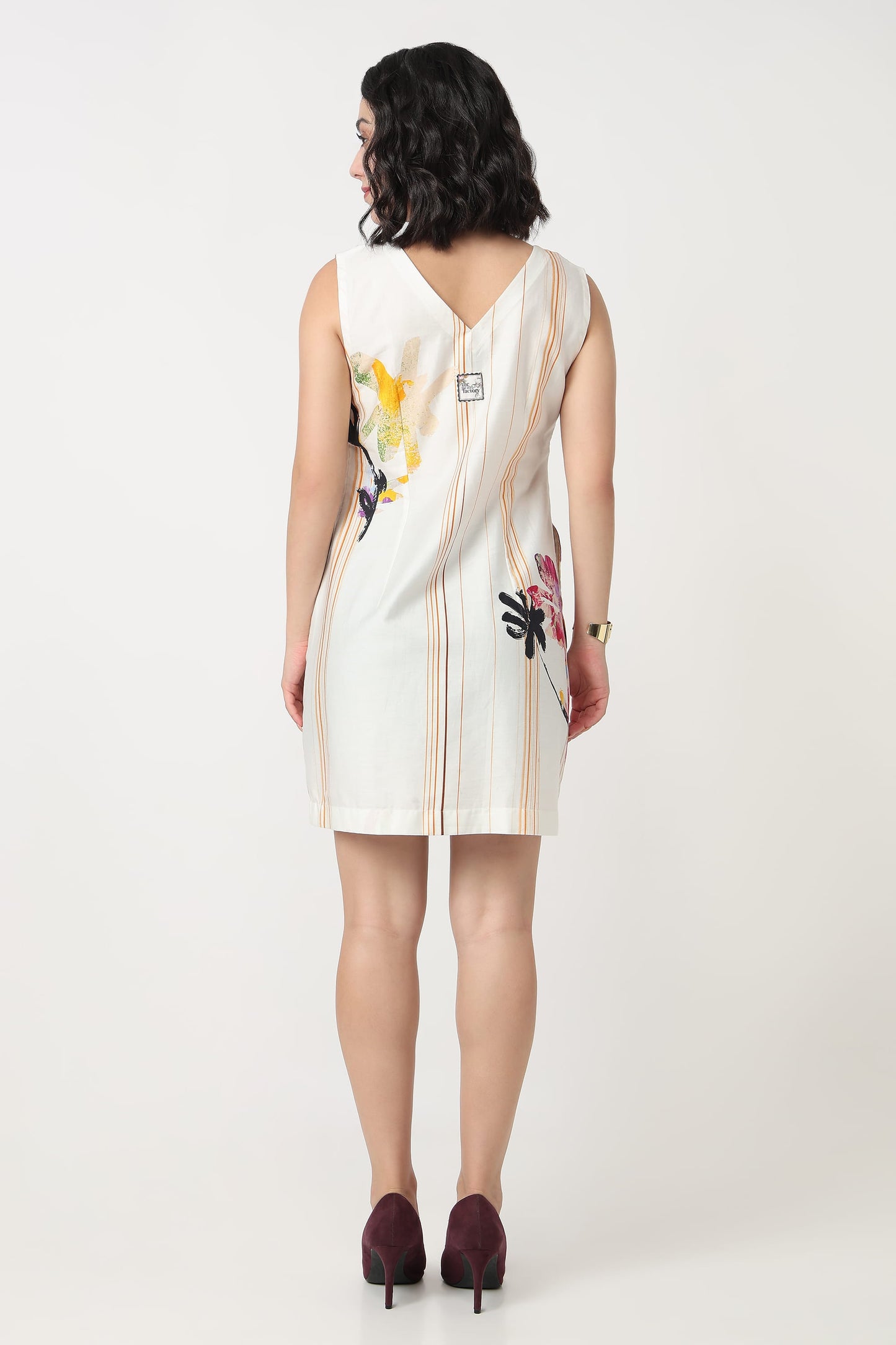 A woman wearing a sleeveless V-neck dress with a colourful floral print and vertical stripes on the bodice and, paired with black heels, against a plain light grey background.