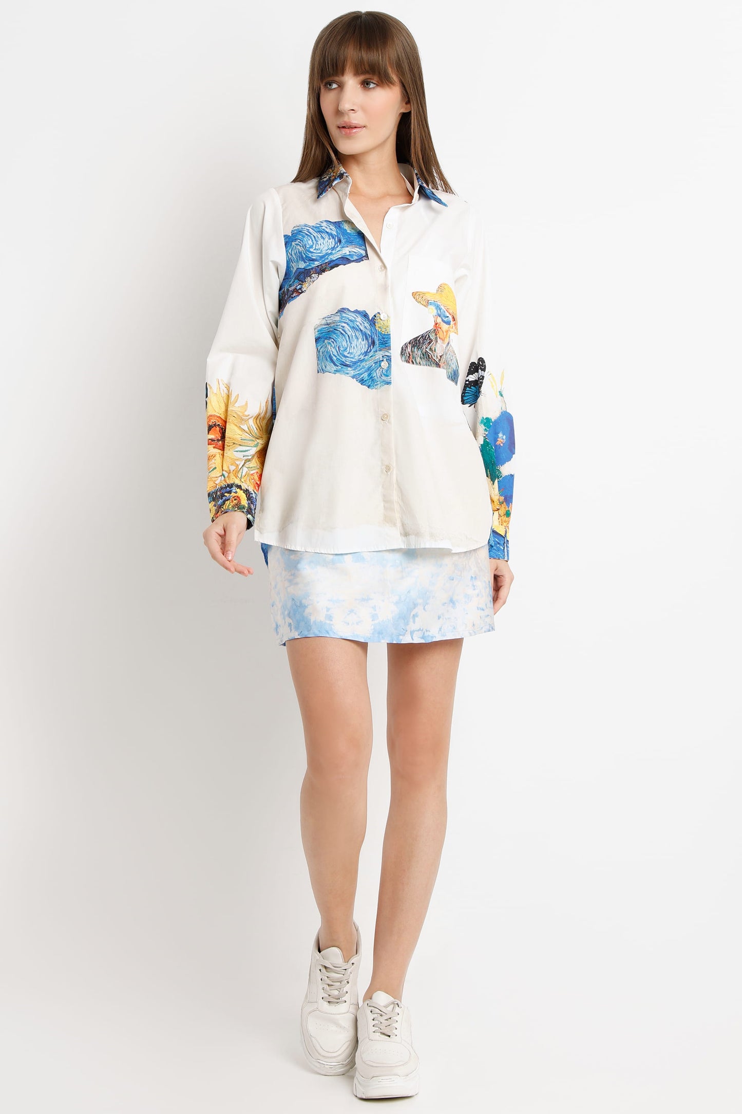 A woman modeling a long-sleeve button-up shirt with a blue and yellow abstract print over an off-white base, paired with a matching light blue patterned mini skirt and white sneakers, against a plain light gray studio background.