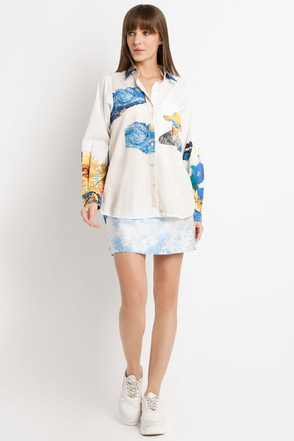 A woman modeling a long-sleeve button-up shirt with a blue and yellow abstract print over an off-white base, paired with a matching light blue patterned mini skirt and white sneakers, against a plain light gray studio background.