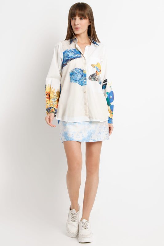 A woman modeling a long-sleeve button-up shirt with a blue and yellow abstract print over an off-white base, paired with a matching light blue patterned mini skirt and white sneakers, against a plain light gray studio background.