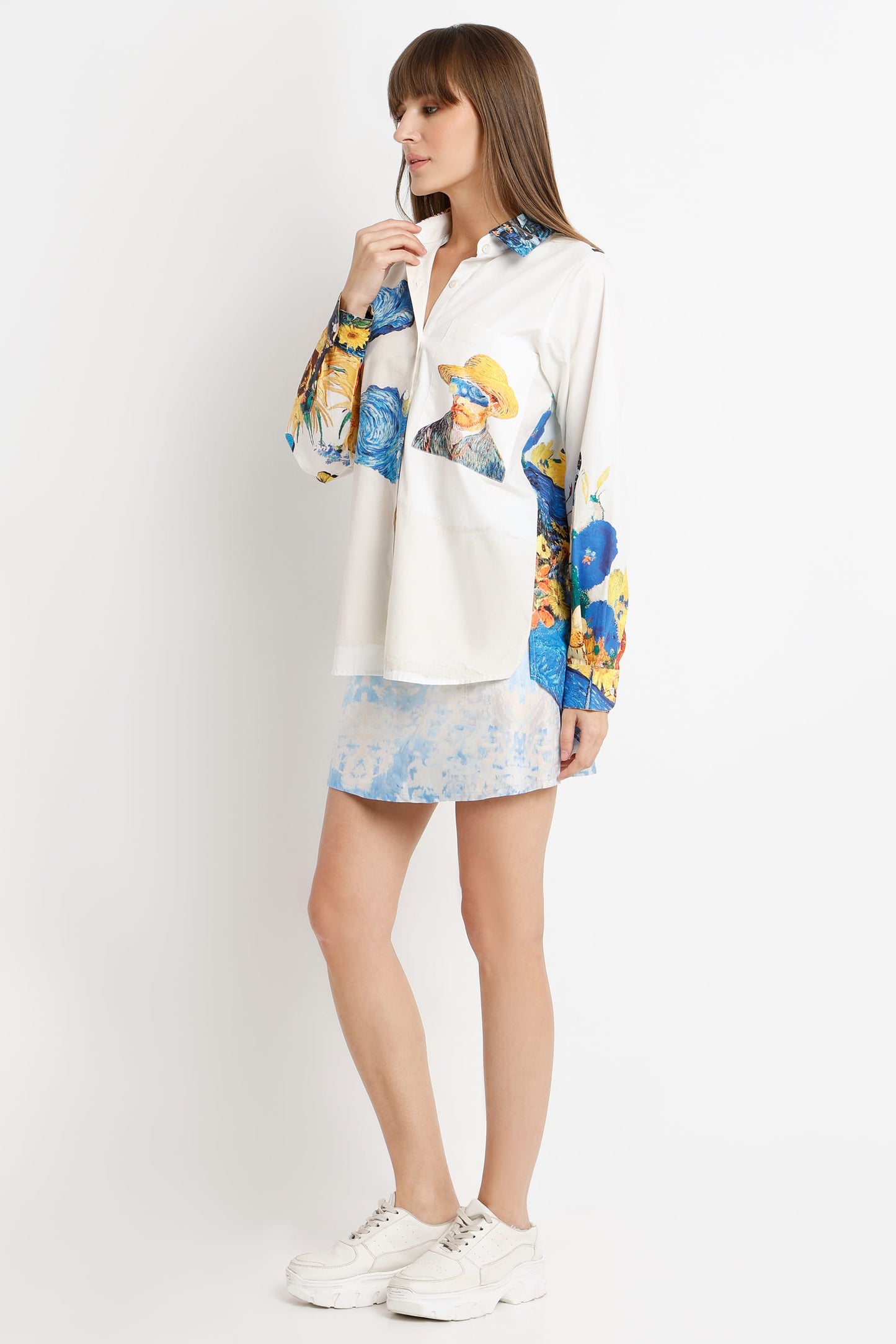 A woman modeling a white long-sleeve shirt with colorful blue, yellow, and green abstract floral patterns on the sleeves and chest, paired with a matching light blue patterned mini skirt and white sneakers, against a plain white studio background.