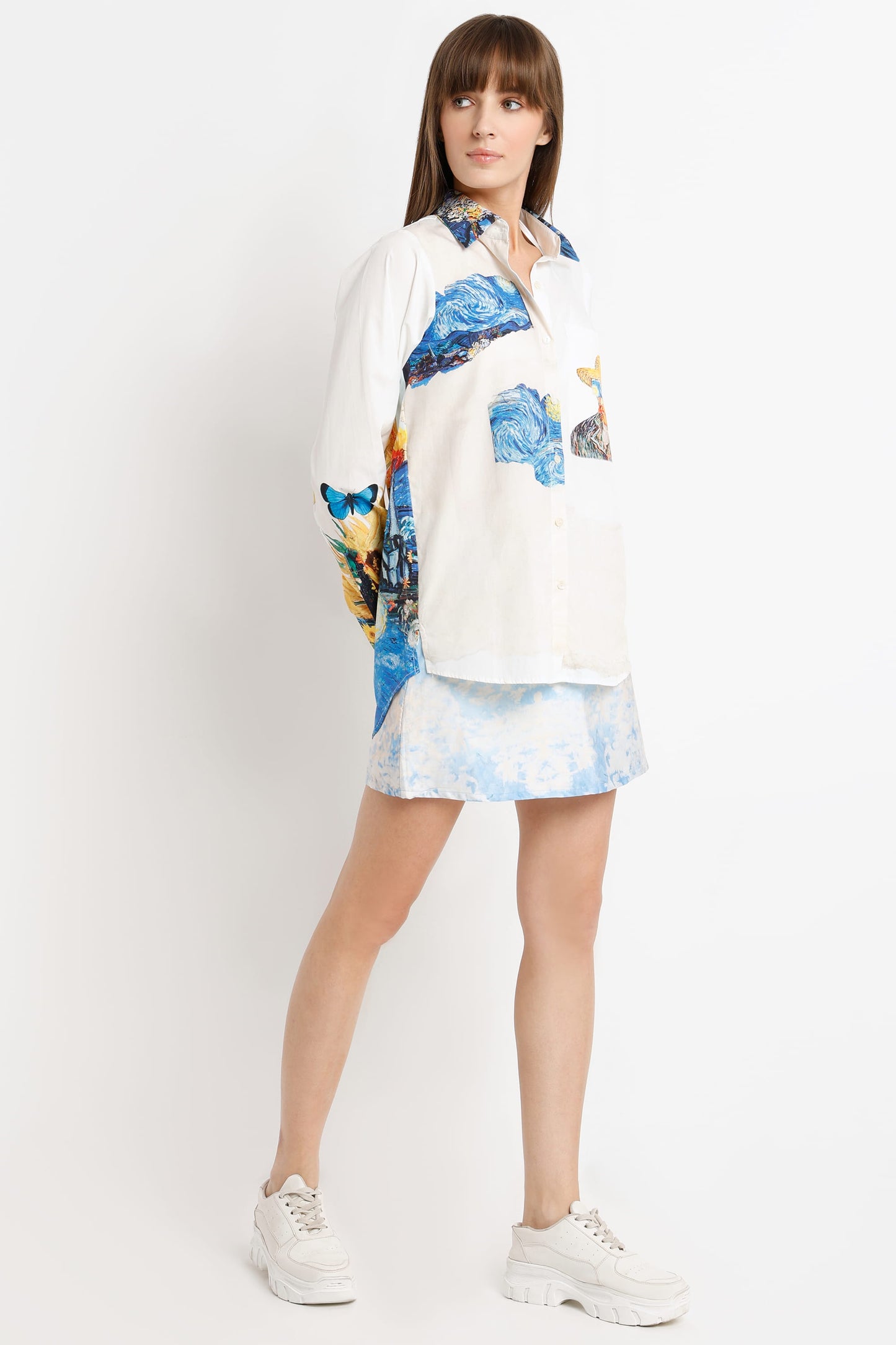 A woman modeling a white button-up shirt with colorful abstract blue and yellow prints, paired with a matching white and blue tie-dye mini skirt and white sneakers, against a plain white studio background.