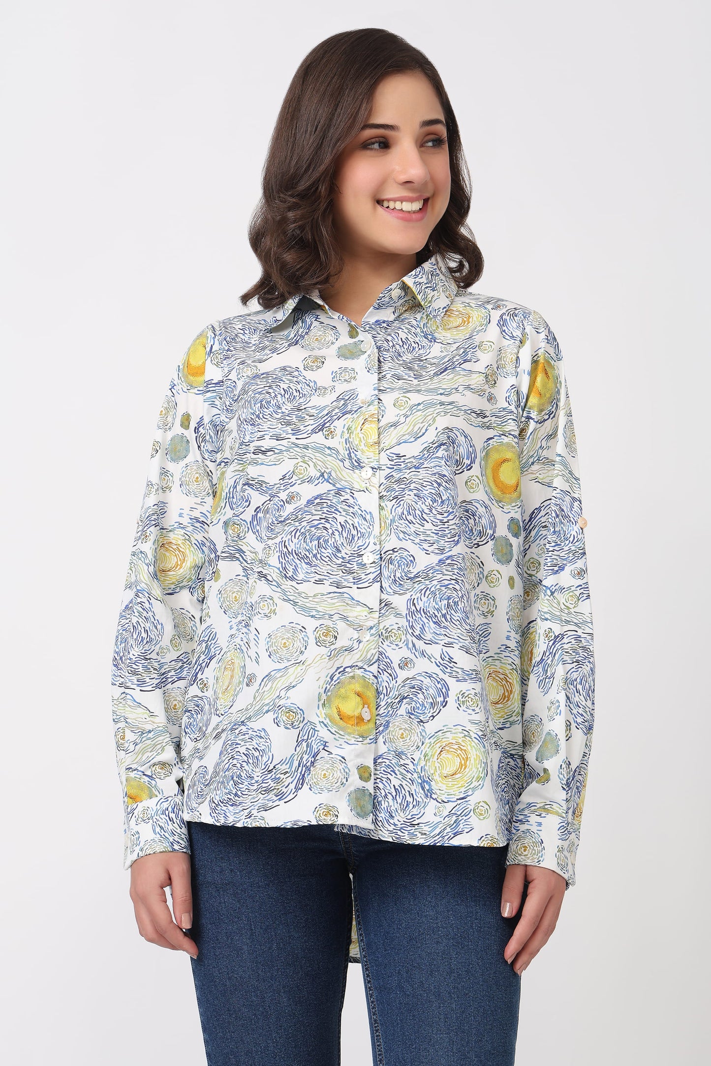 A woman wearing a long-sleeve button-up shirt with a blue and yellow floral pattern on a white background, paired with dark blue jeans, against a light gray studio background.