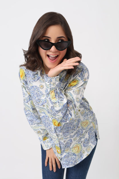 A woman wearing a long-sleeve button-up shirt with a blue and yellow floral pattern, styled with dark sunglasses and blue jeans against a plain white background.