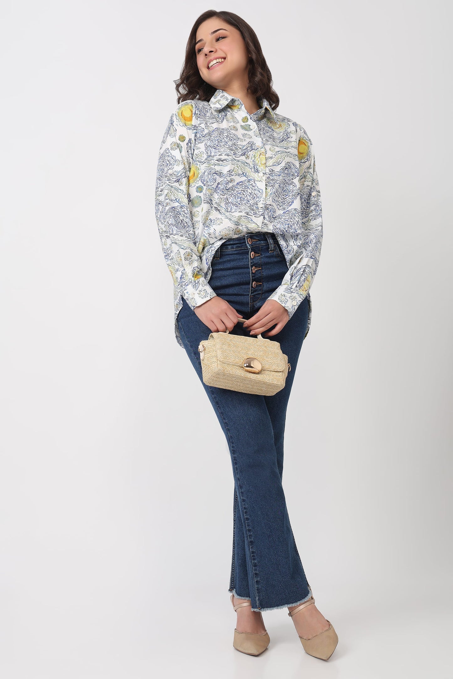 A woman modeling a long-sleeve button-up shirt with a blue and yellow toile pattern, paired with dark blue jeans, beige heels, and holding a beige handbag against a light gray studio background.