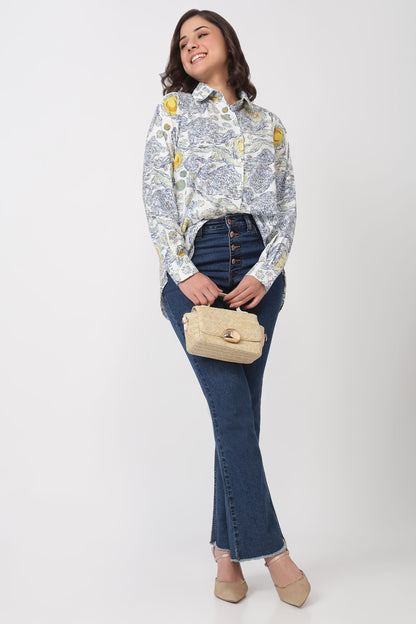 A woman modeling a long-sleeve button-up shirt with a blue and yellow toile pattern, paired with dark blue jeans, beige heels, and holding a beige handbag against a light gray studio background.